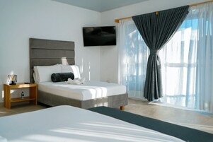 A modern hotel room with a large bed, a bedside table with a lamp, a flat-screen TV mounted on the wall, and a large window with sheer curtains.