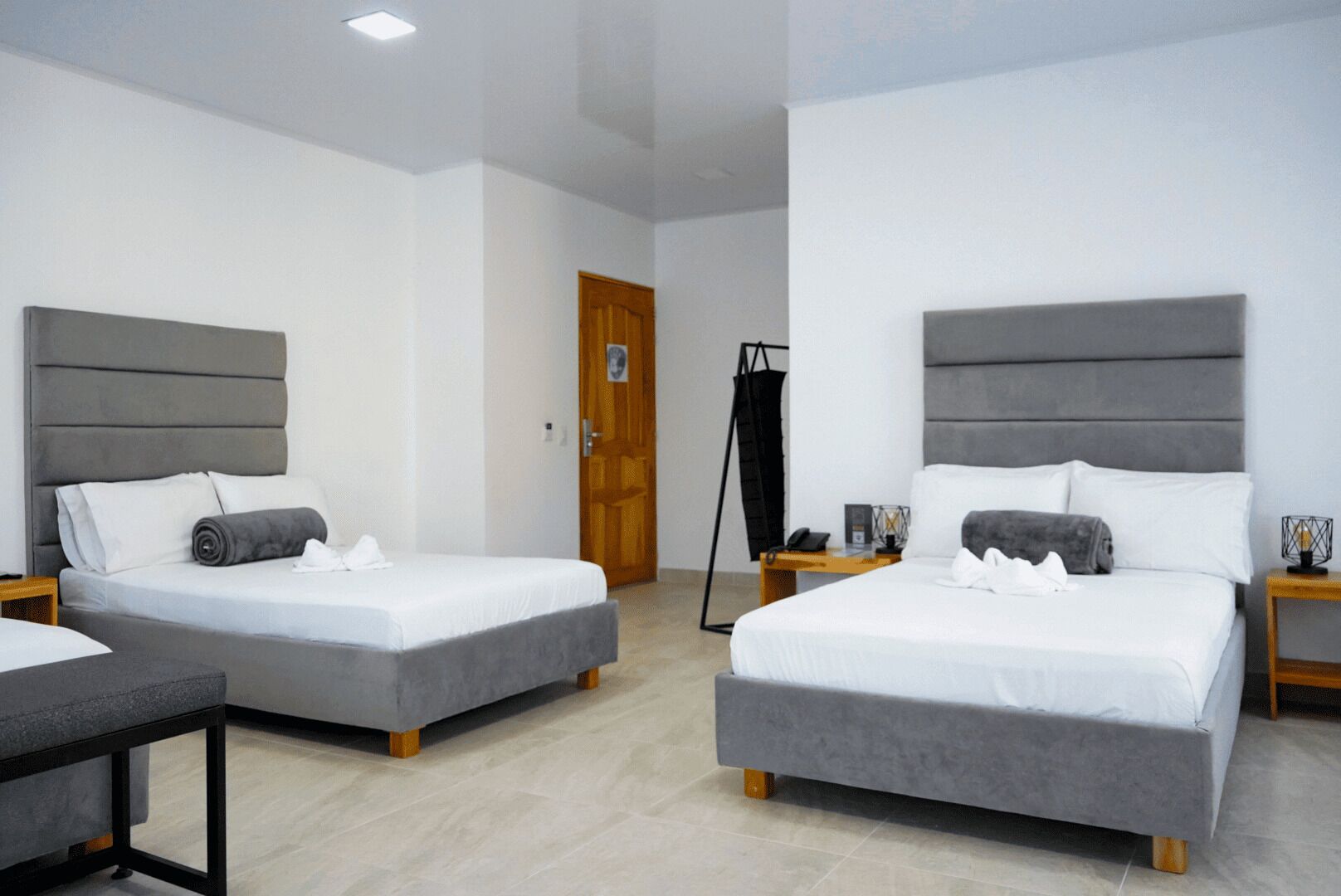 A hotel room with two beds, each with a grey headboard and white bedding. There is a wooden door in the background, a black coat rack, and a small table with a lamp and a picture frame.