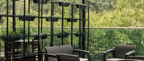 Een terras buiten met rieten meubilair en een zwart metalen trellis met hangplanten.