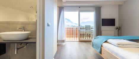 Ett modernt hotellrum med ett stort handfat på badrummet, en säng med ett blått överkast, en väggmonterad TV och en balkong med utsikt över bergen.