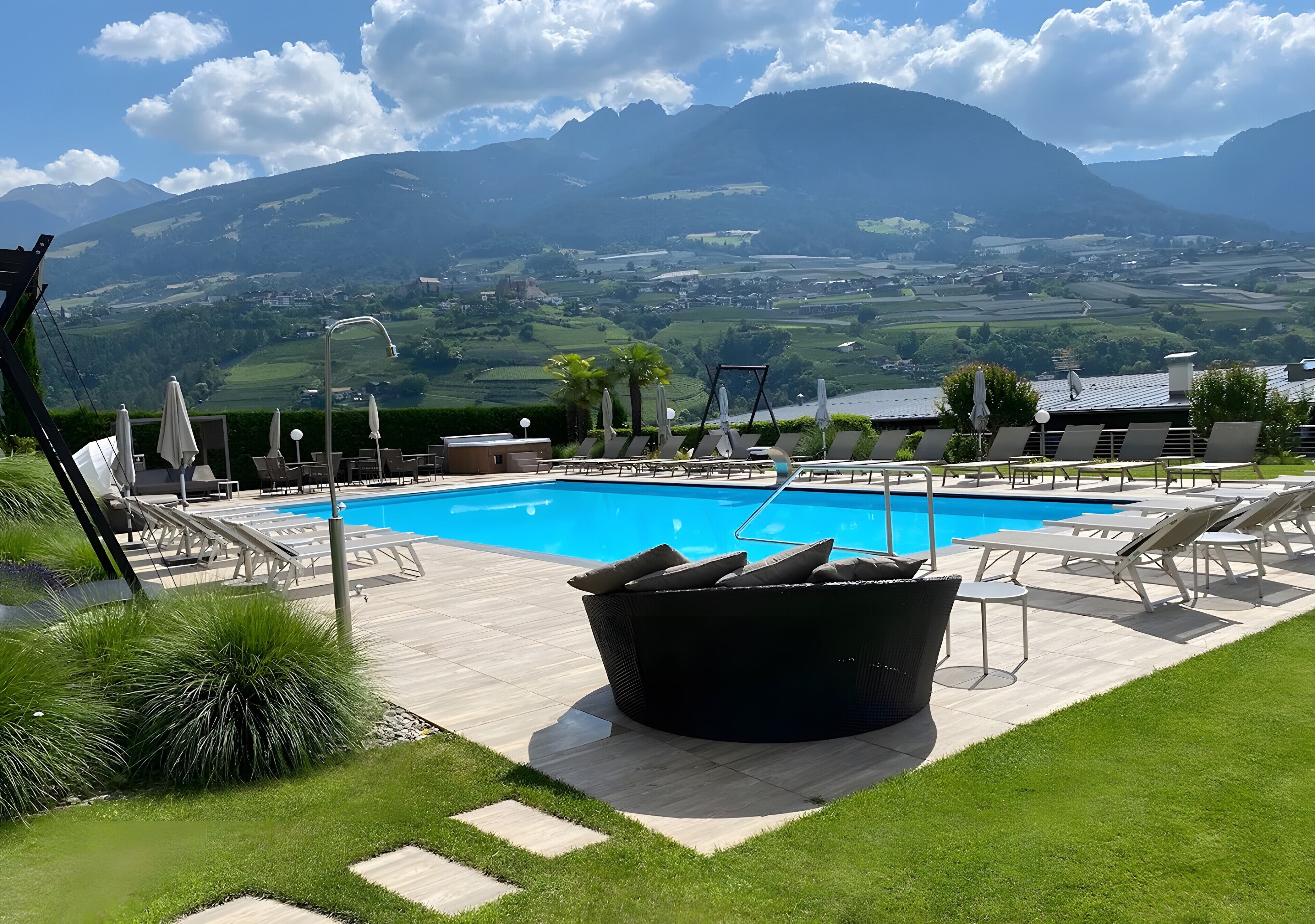 A pool area with lounge chairs and a hot tub, surrounded by a lawn and mountains in the background.