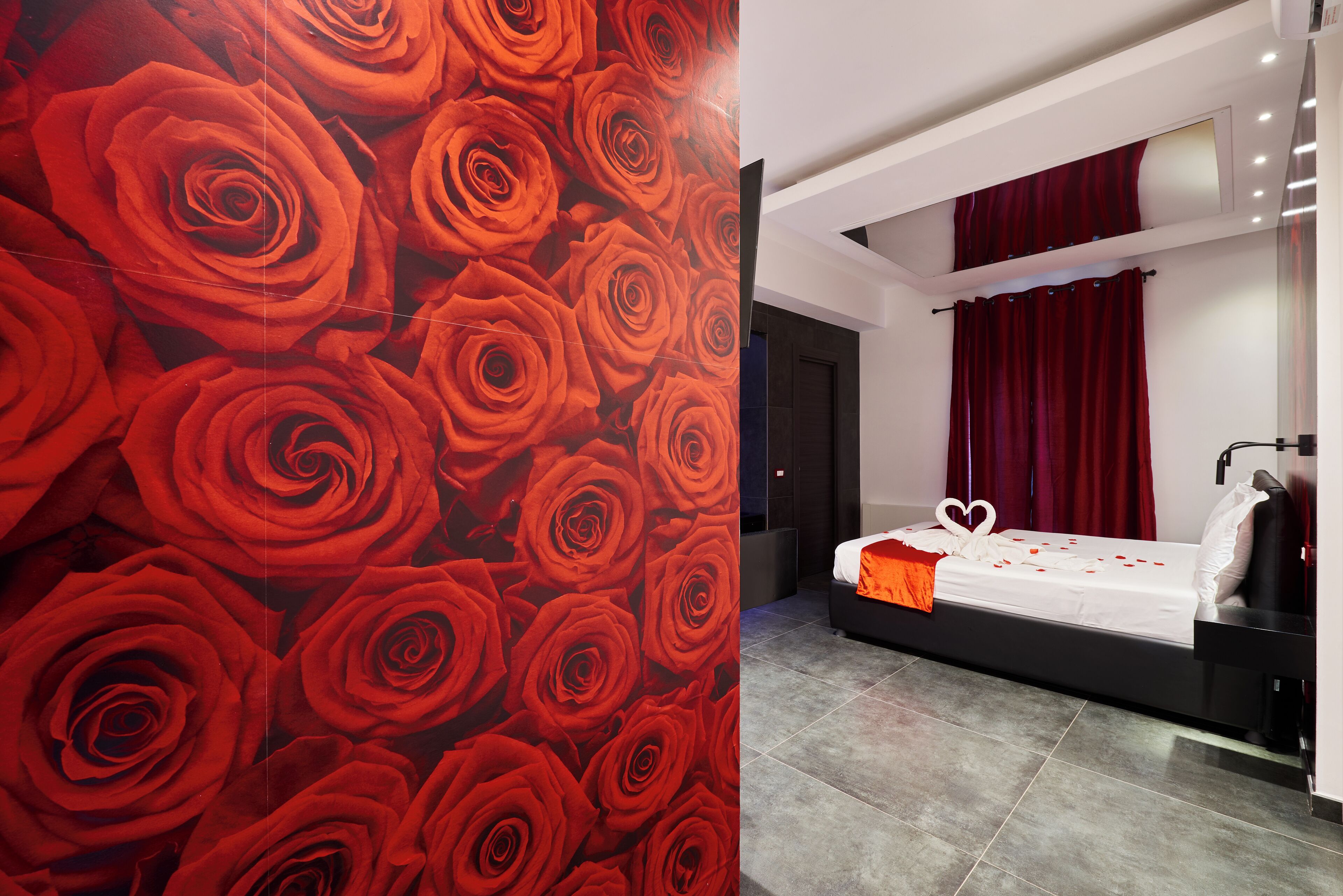 A modern hotel room with a large wall mural of red roses, a neatly made bed with a heart-shaped pillow, and a red curtain.
