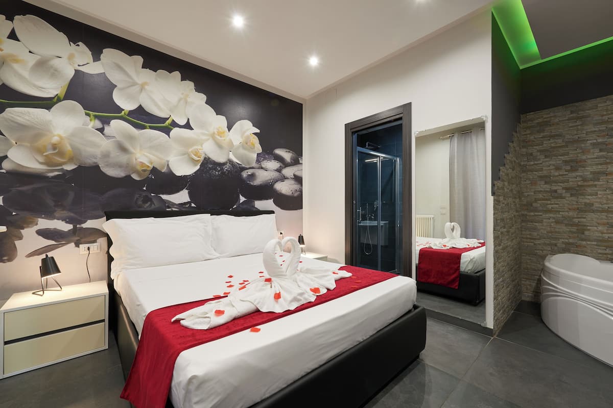 A modern bedroom with a large bed, a nightstand, a mirror, a stone wall, and a bathroom with a bathtub.