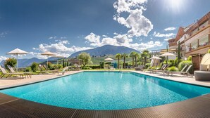 A rectangular swimming pool with lounge chairs and umbrellas, surrounded by a mountainous landscape and a multi-story building with balconies. - Room 'Silber Zimmer 2' with Mountain View, Wi-Fi and Air Conditioning (Tirolo)