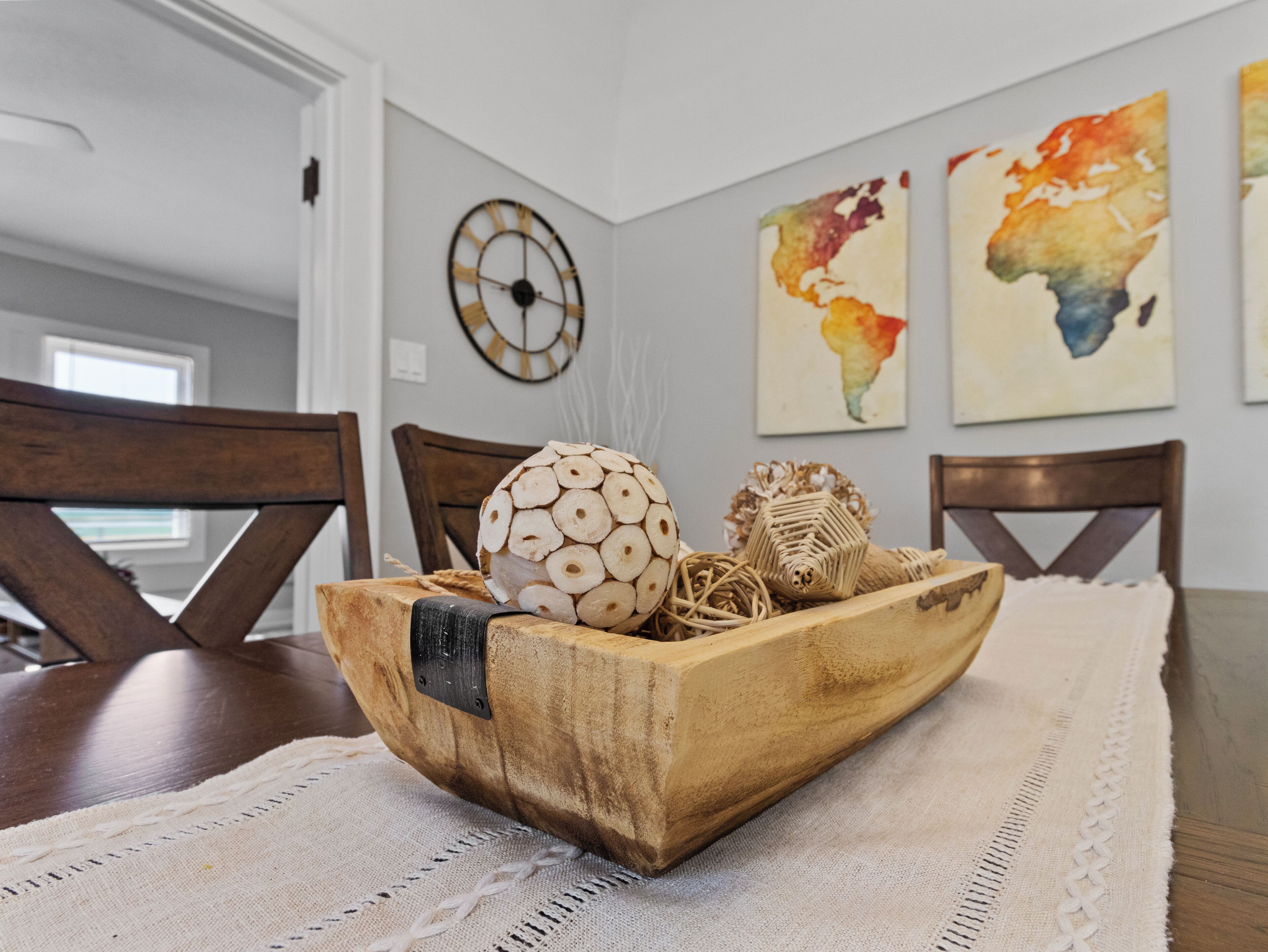 Un bol en bois, orné de divers objets décoratifs, est posé sur une table devant un mur où sont accrochés une horloge et deux tableaux abstraits.