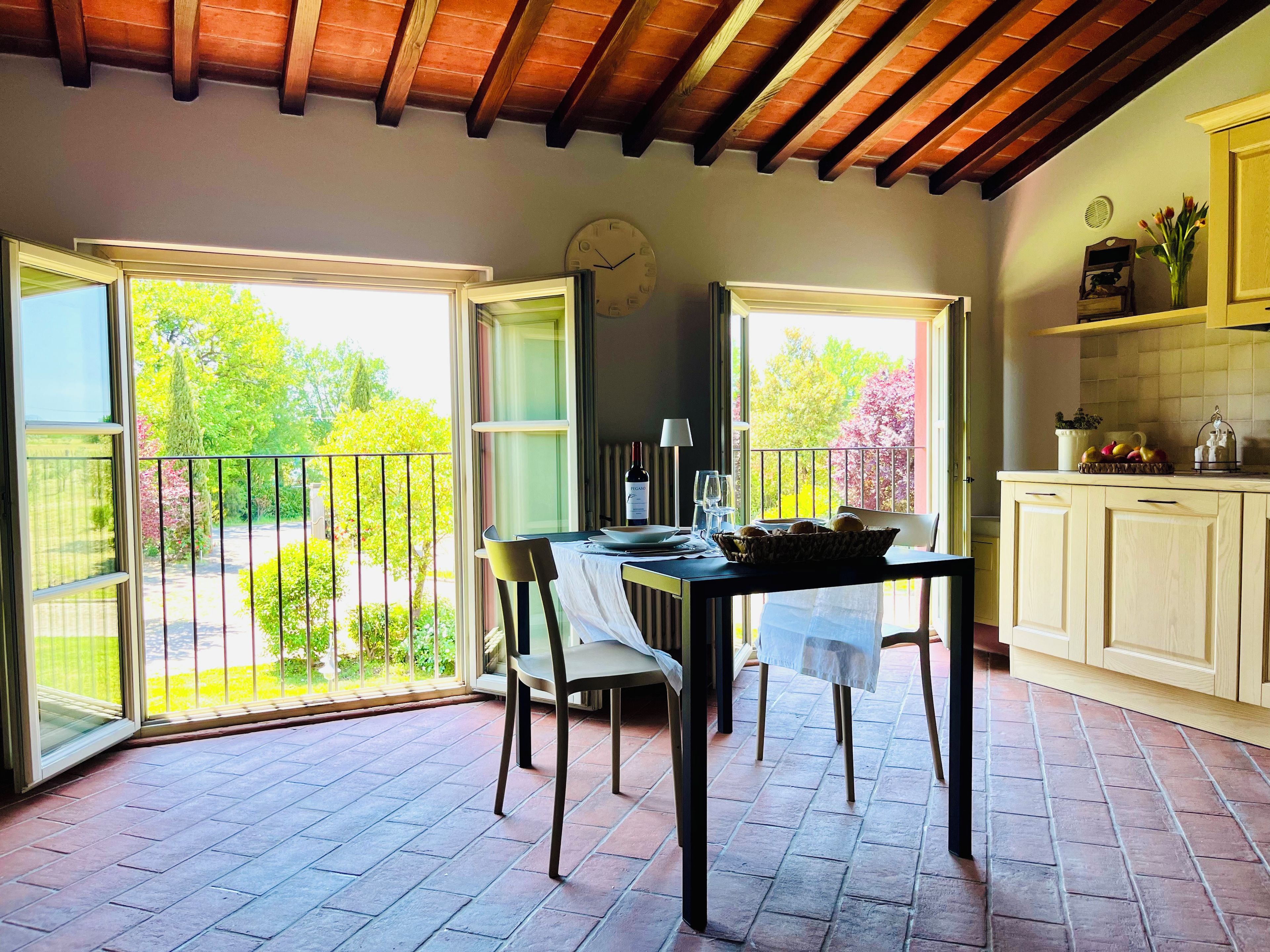 Un coin repas avec une table dressée pour deux personnes, une horloge au mur et une vue sur le jardin par des portes ouvertes.