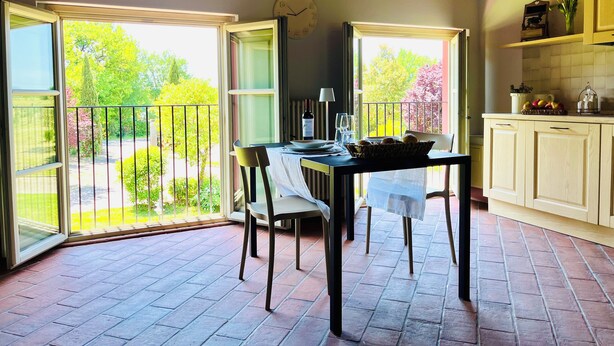 Uma sala de jantar com uma mesa posta para dois, um relógio na parede e vista para o jardim através de portas abertas.