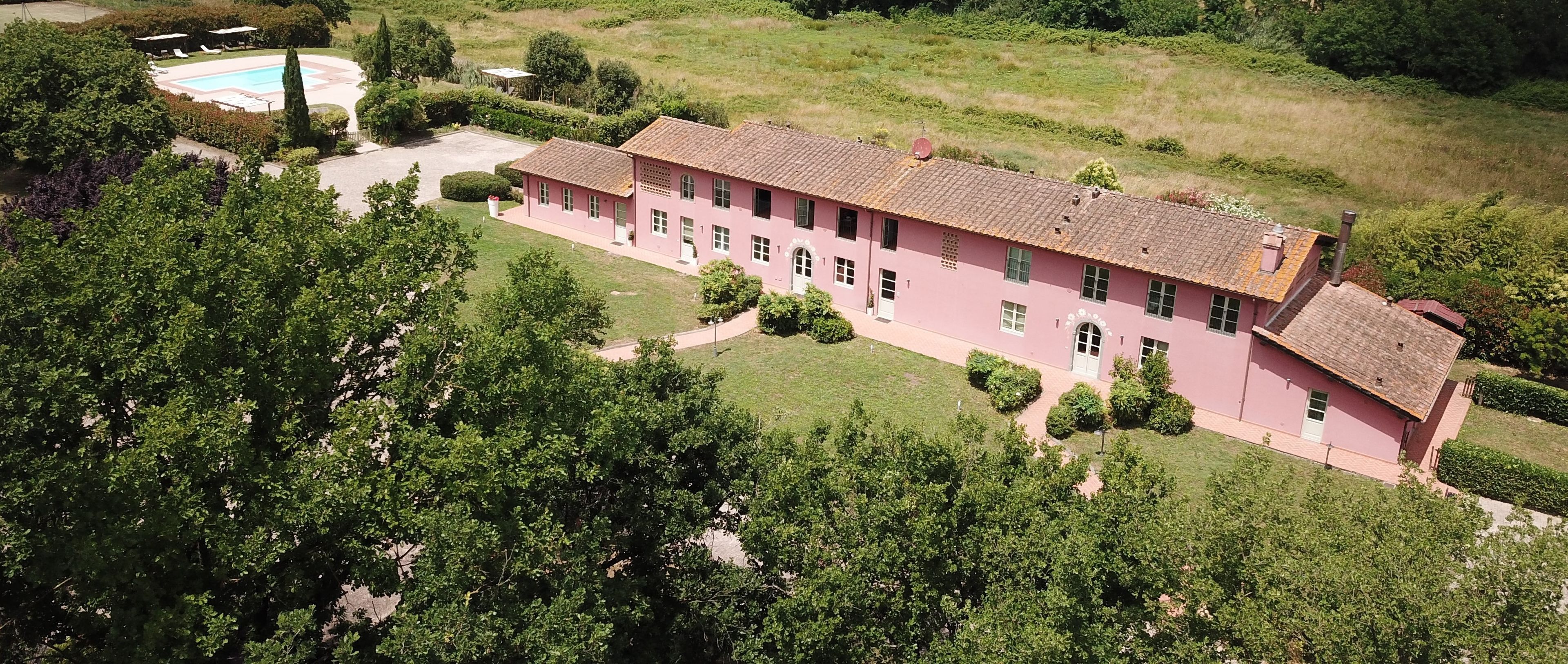Un bâtiment rose avec un toit en tuiles, entouré de verdure et agrémenté d’une piscine.