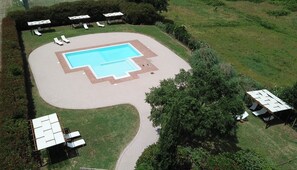 An aerial view of a swimming pool area with a surrounding deck, lounge chairs, and a paved walkway.