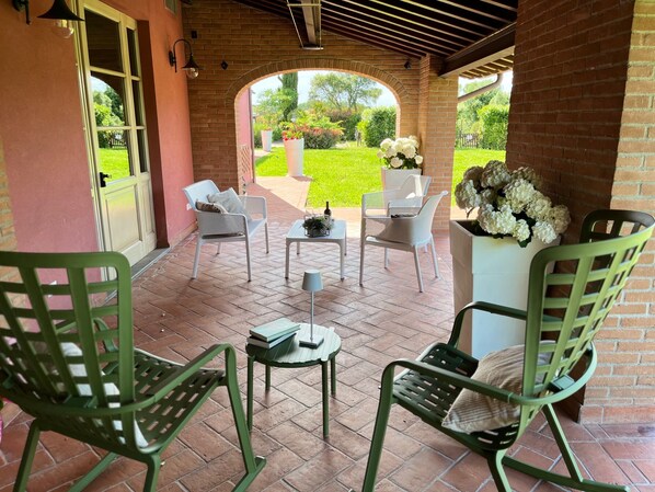 An outdoor patio with brick flooring, white wicker chairs, a small round table with books and a lamp, and a flower arrangement.