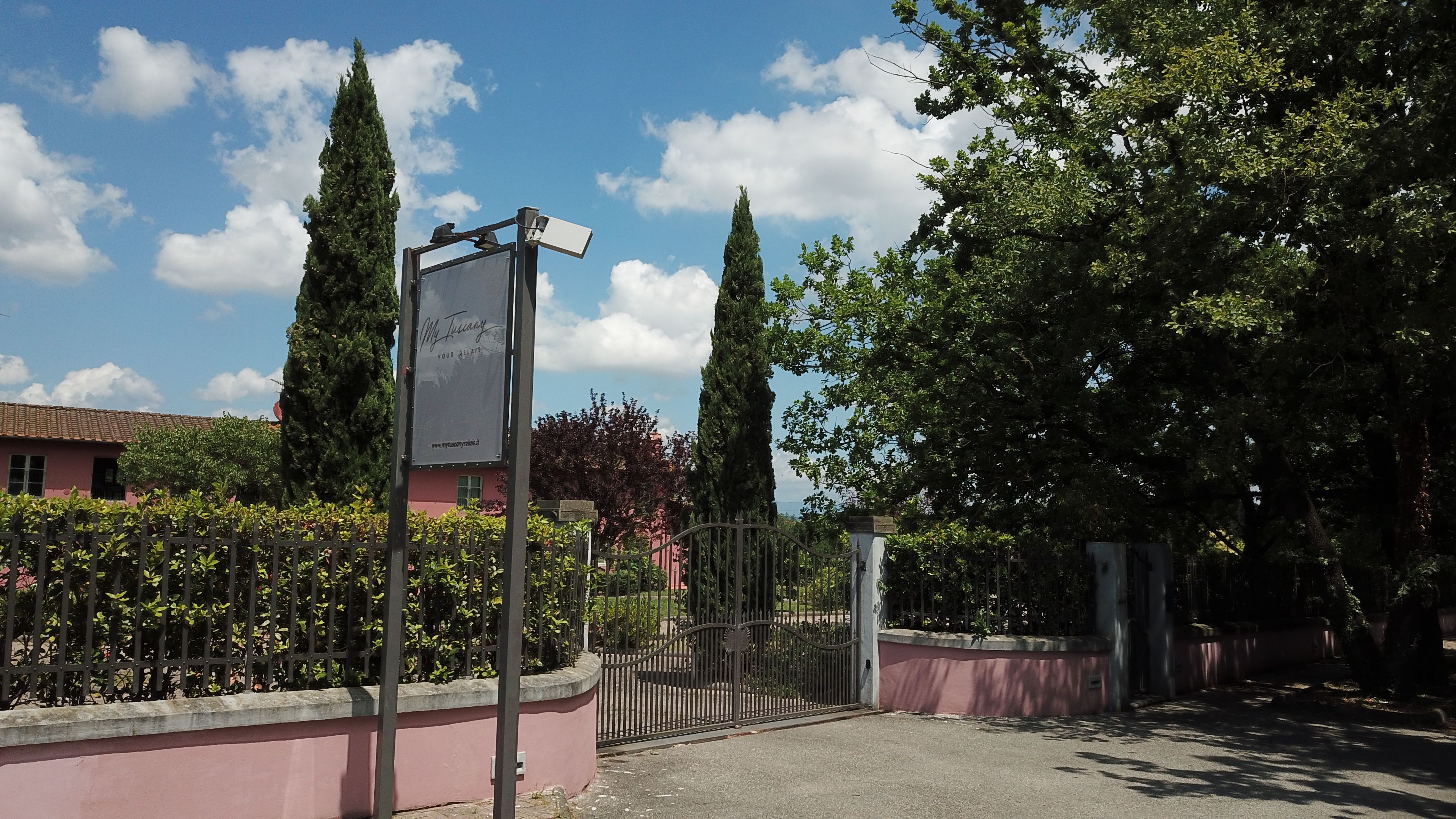 A gated entrance with a sign, a pink building, and tall trees.