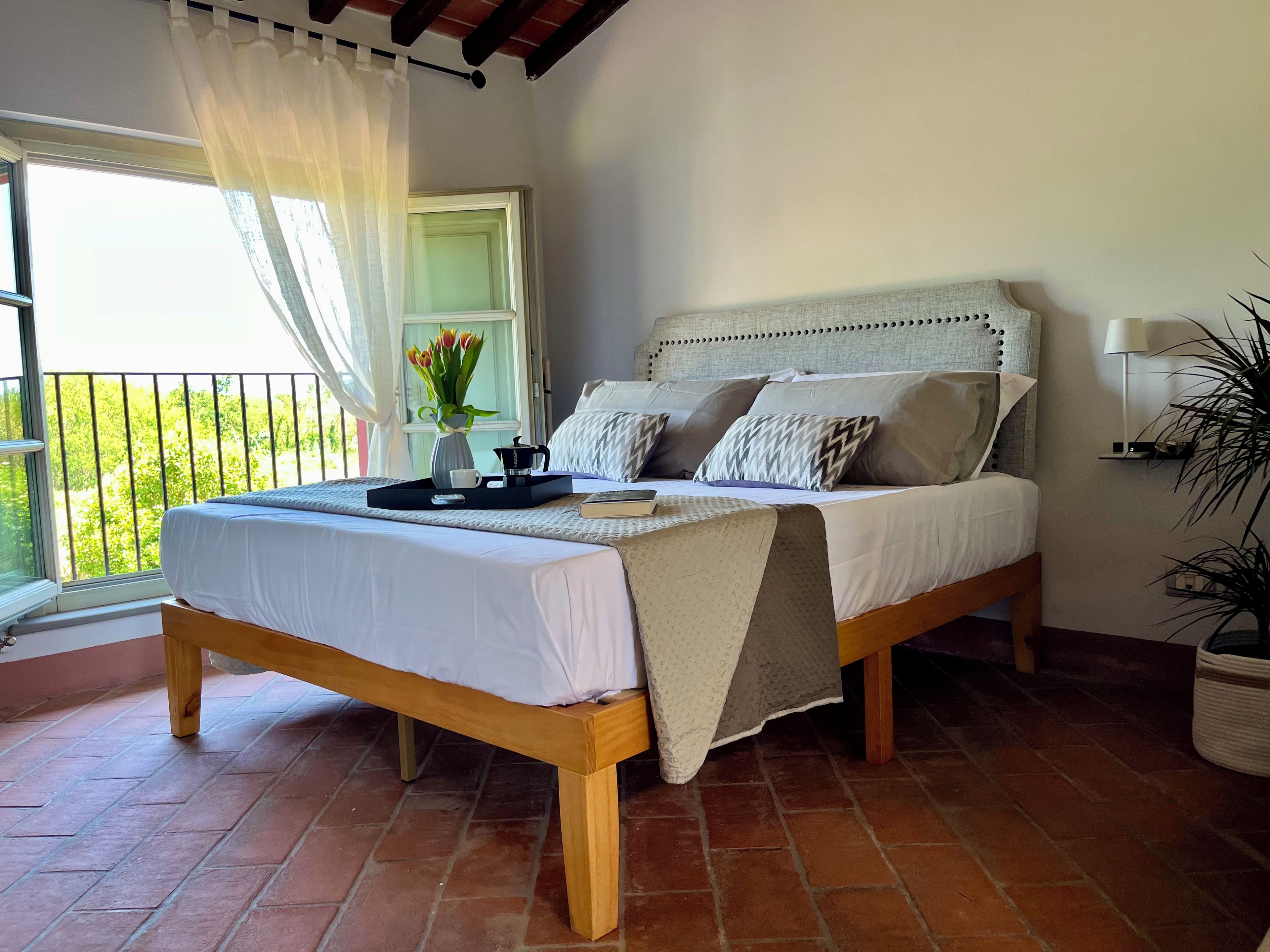 Un letto con testiera, un comodino, una finestra con tende e vista sul verde circostante.