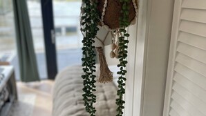 Un tapiz de macramé con follaje verde y adornos esféricos, colgado cerca de una ventana con persianas.