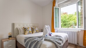 Una cama bien hecha con una manta gris, almohadas blancas y un cabecero blanco. Hay una mesita de noche de madera con una lámpara y un globo terráqueo. Se ve una ventana con cortinas amarillas, y un radiador debajo de la ventana.