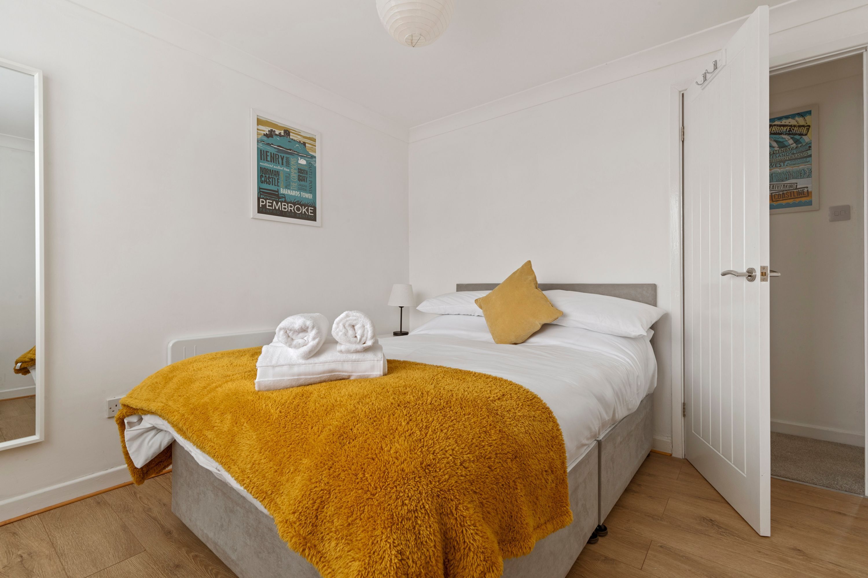A bedroom with a bed, a yellow blanket, two towels, a pillow, a poster on the wall, and a door.