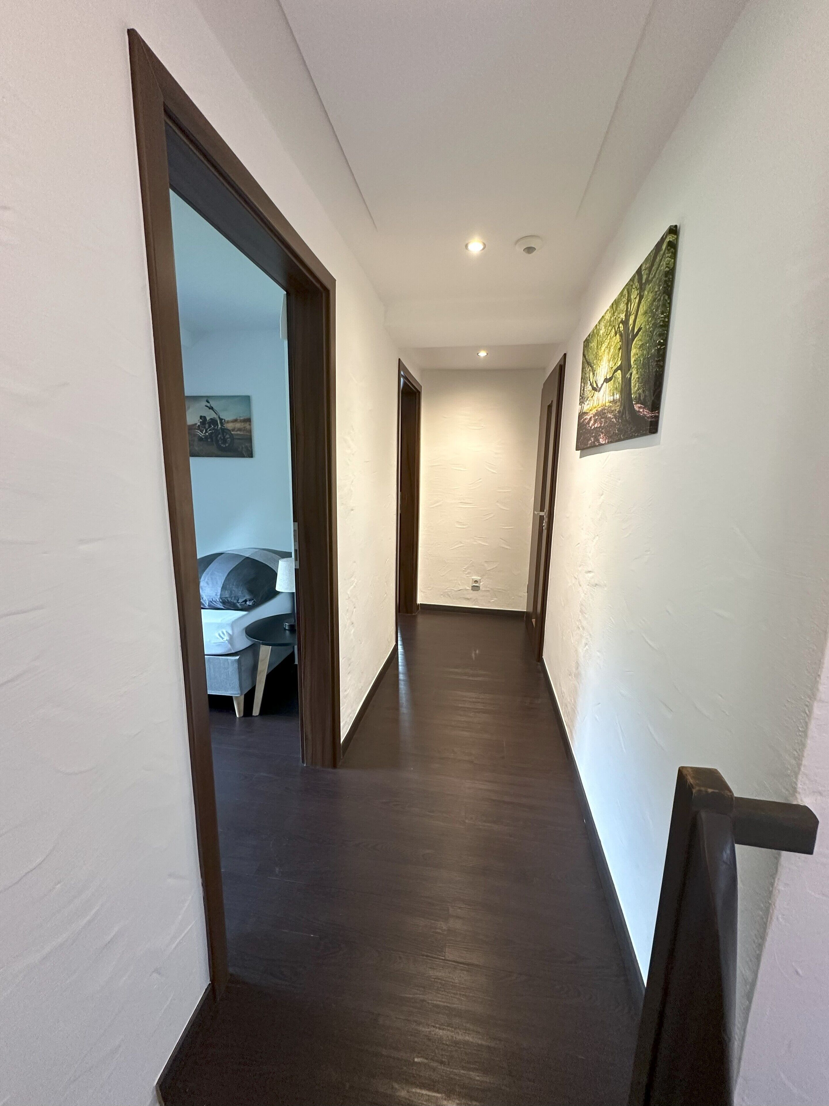 Ein schmaler Flur mit dunklem Holzboden, weißen Wänden und einer Decke mit eingelassener Beleuchtung. In der linken Wand befindet sich eine Tür, die zu einem Zimmer mit Bett und Stuhl führt. Auf der rechten Wand ist ein Gemälde eines Waldes zu sehen.