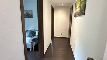 Ein schmaler Flur mit dunklem Holzboden, weißen Wänden und einer Decke mit eingelassener Beleuchtung. In der linken Wand befindet sich eine Tür, die zu einem Zimmer mit Bett und Stuhl führt. Auf der rechten Wand ist ein Gemälde eines Waldes zu sehen.