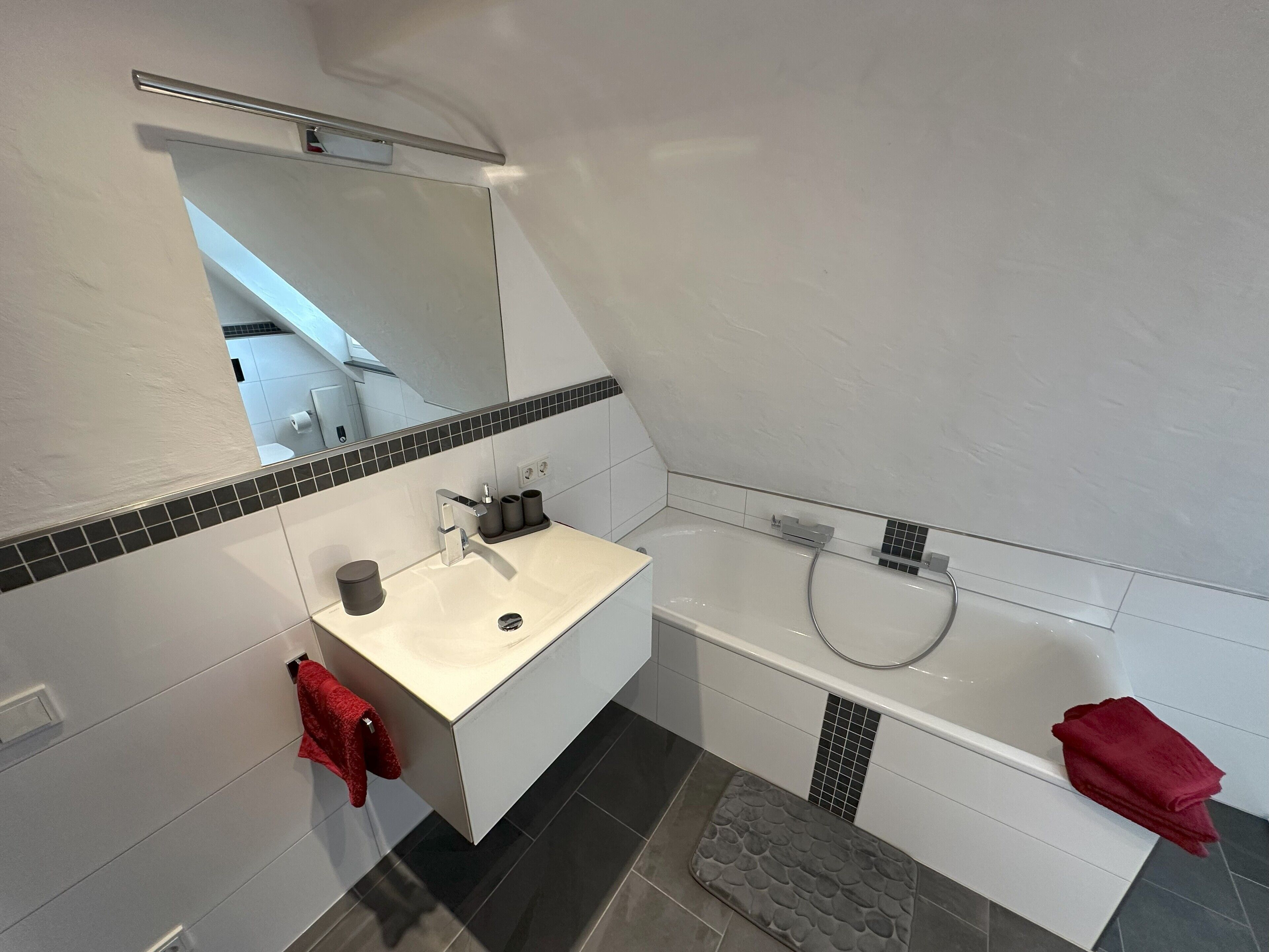 A modern bathroom with a white vanity, a large mirror, a bathtub, and a red towel.
