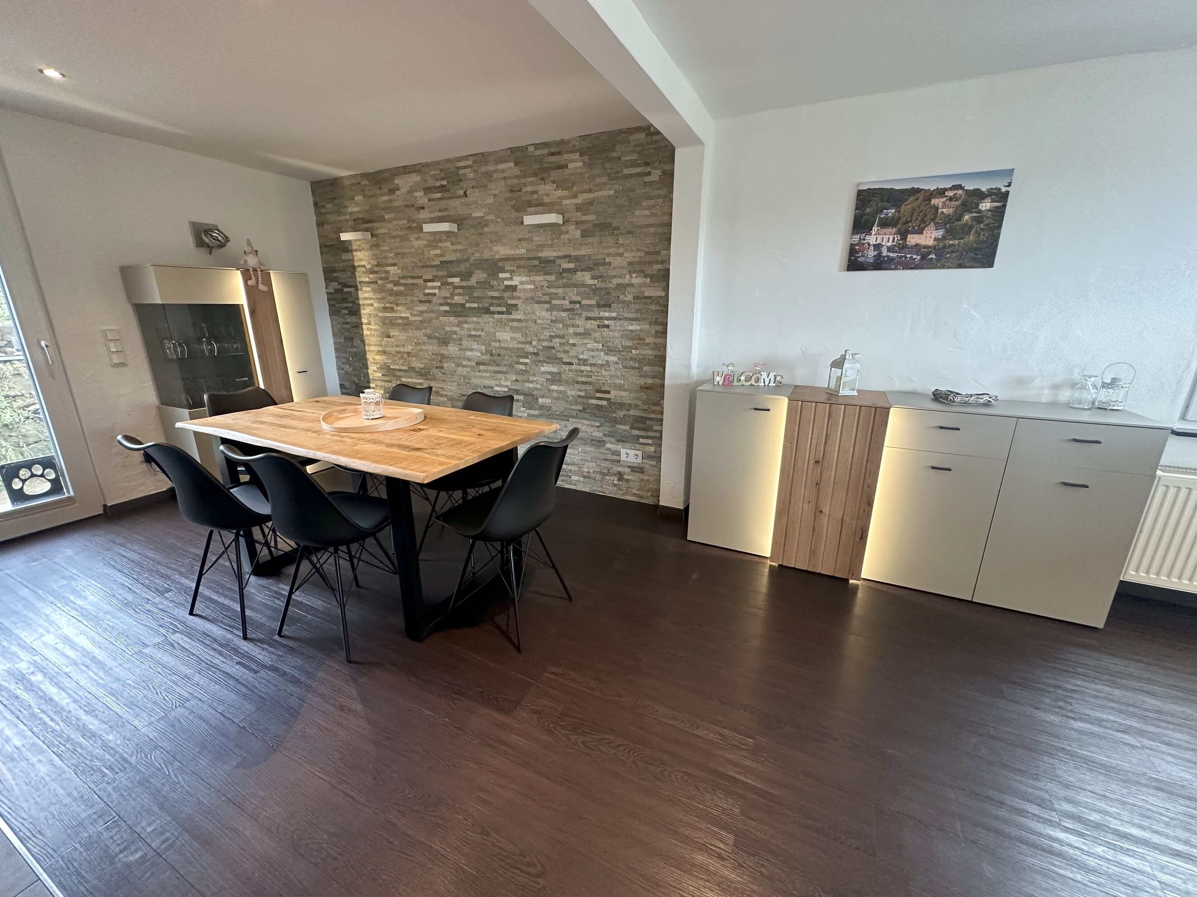 Une cuisine moderne avec un mur arrière en pierre, des armoires en bois et un coin repas comprenant une table en bois et des chaises noires.