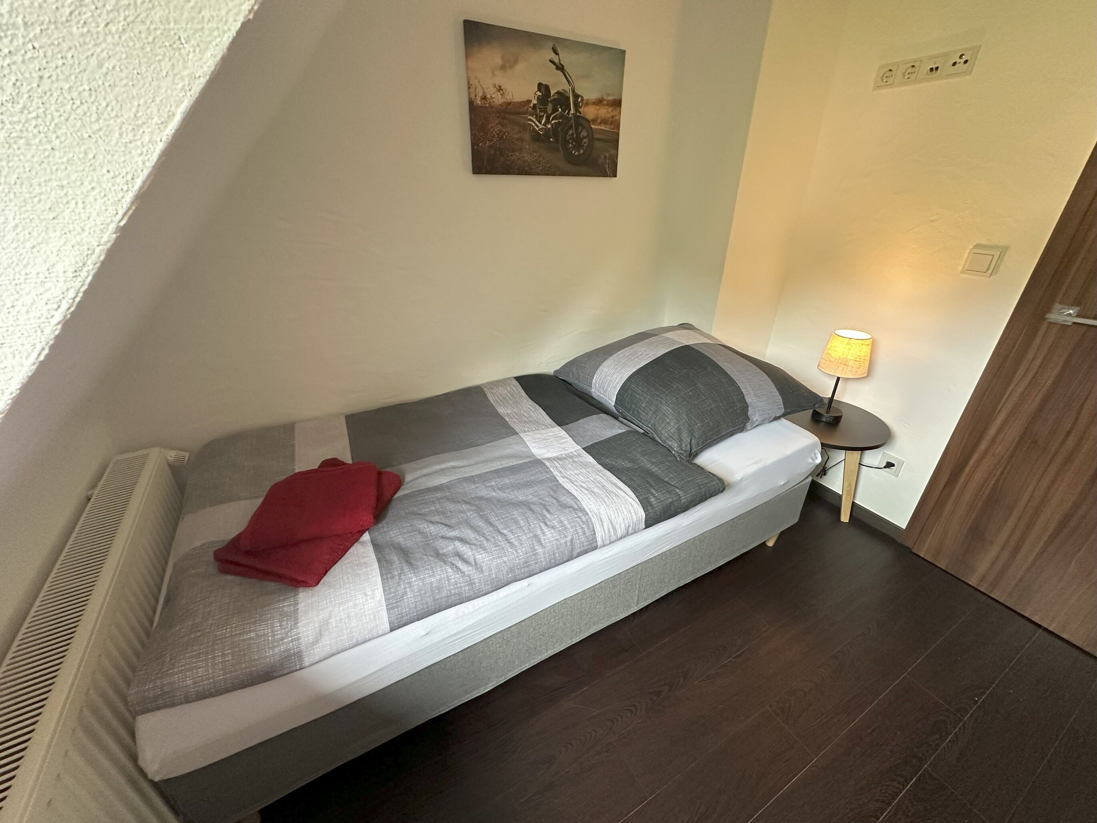 Una habitación pequeña con una cama individual, cubierta con un edredón a rayas gris y blanco, una manta roja, un cuadro en la pared de una motocicleta, una mesita de noche con una lámpara y una puerta de madera.