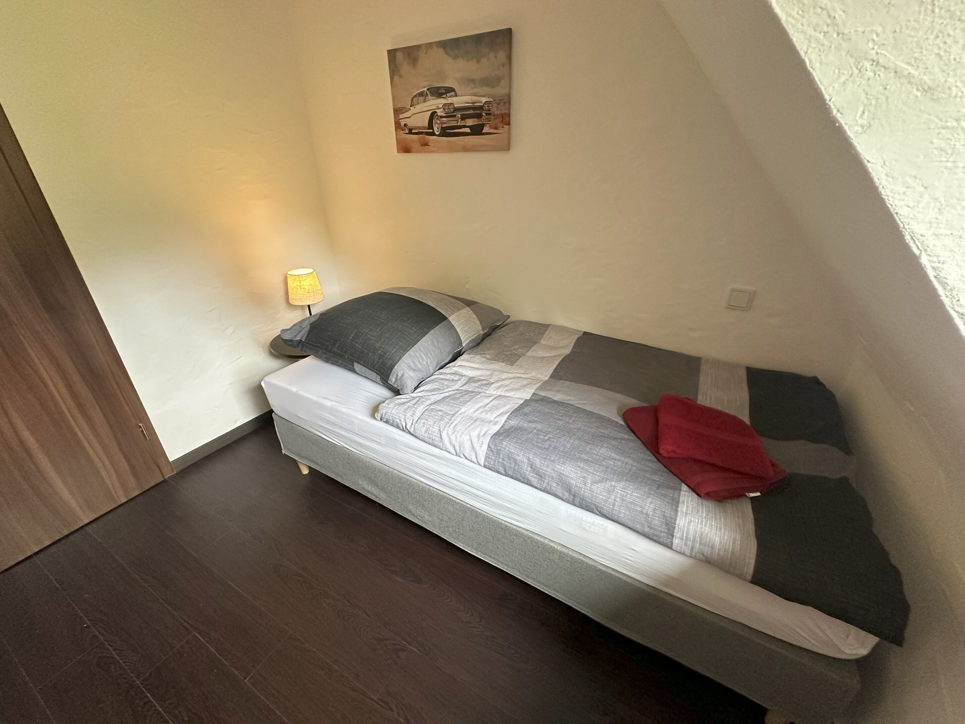 Een kleine kamer met een enkel bed, een houten vloer, een bedlampje, een schilderij aan de muur en een rode handdoek op het bed.