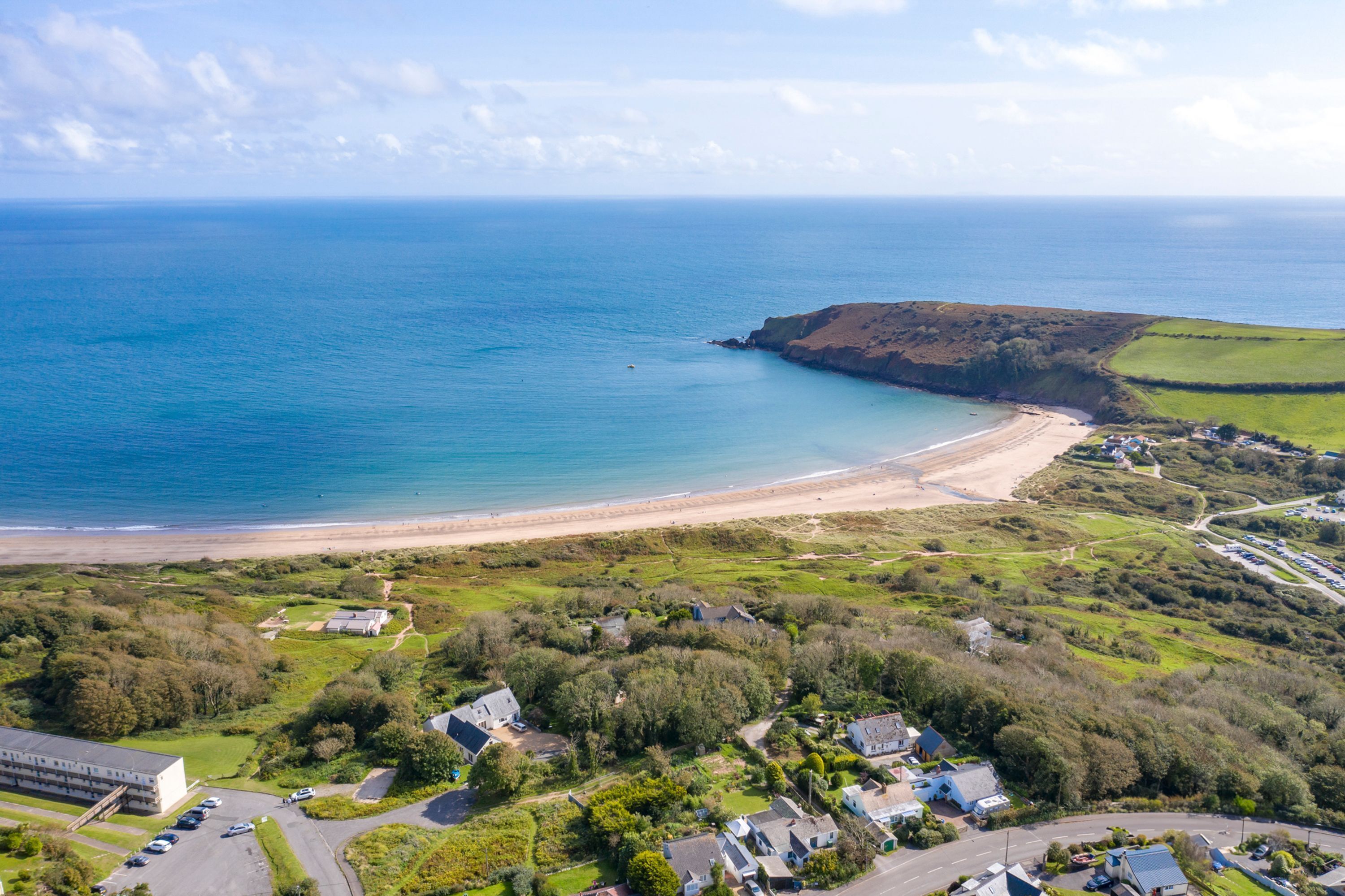 An aerial view of a coastal area with a beach, green fields, and residential buildings.