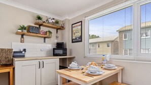 A kitchen with white cabinets, a wooden table, and a window with blinds. There are plants on shelves and a sign on the wall. - The Retreat - Short walk from beach, parking (Freshwater East)