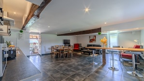 A modern kitchen with dark tiles, a wooden ceiling beam, a dining area with a long table and chairs, and a living room with a red sofa and a TV. - Les Coquelicots - A 30 min de Roanne (Régny)
