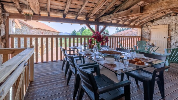 An outdoor dining area with a wooden deck, a table set for two, and a stone wall in the background. - Les Coquelicots - A 30 min de Roanne (Régny)