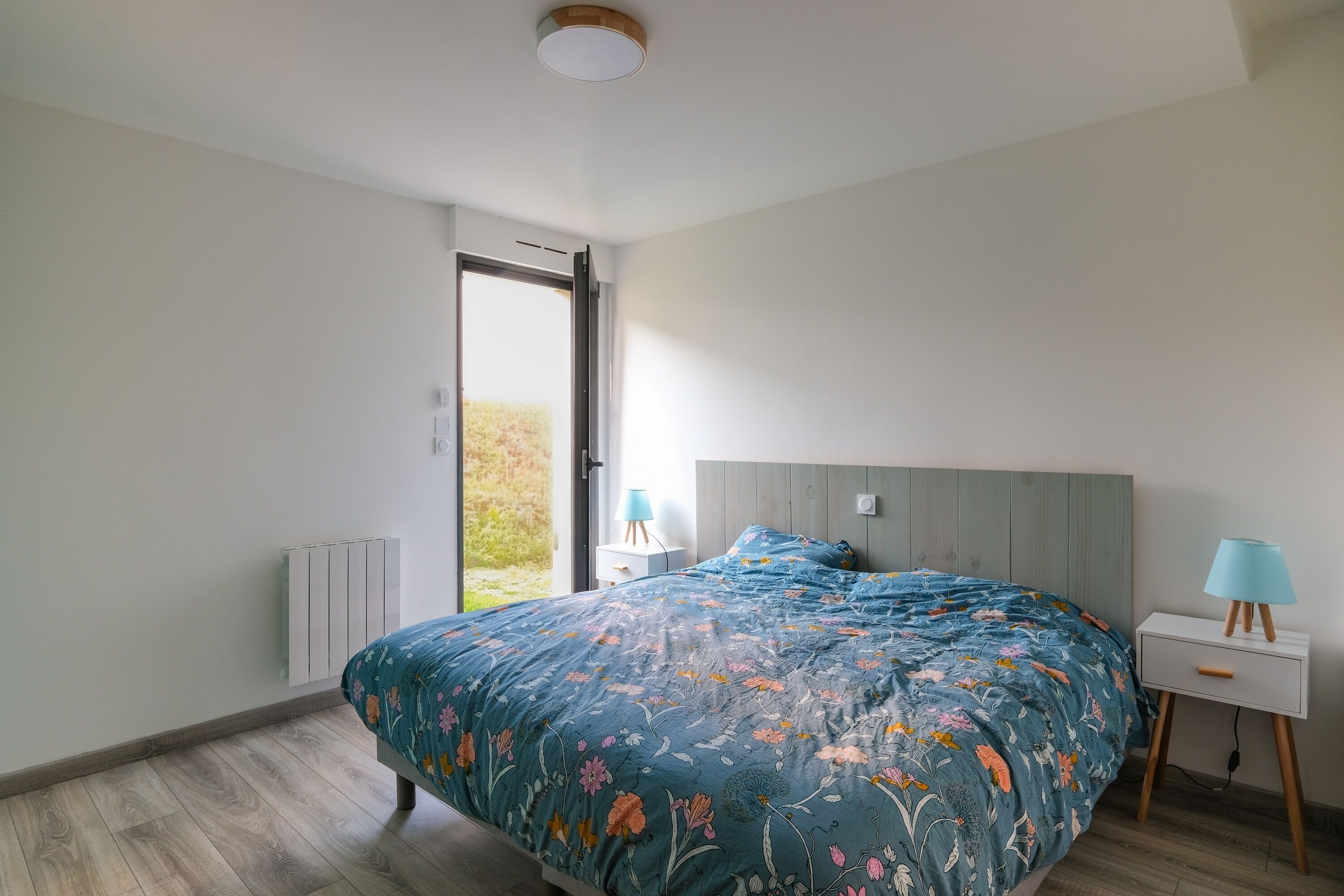 A bedroom with a large bed, a bedside table with a lamp, and a view of the outdoors.