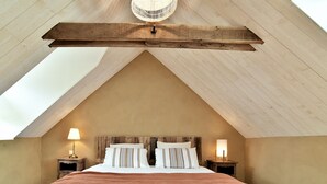 A bedroom with a sloped ceiling, a large bed with white linens and striped pillows, two bedside tables with lamps, and a wooden beam structure.