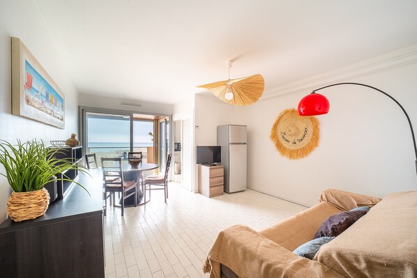 A modern living room with a sofa, a dining area with a table and chairs, a kitchen area with a fridge and a TV, and a large window offering a view of the beach. - Côte Vermeille - Vue mer (Canet-en-Roussillon)