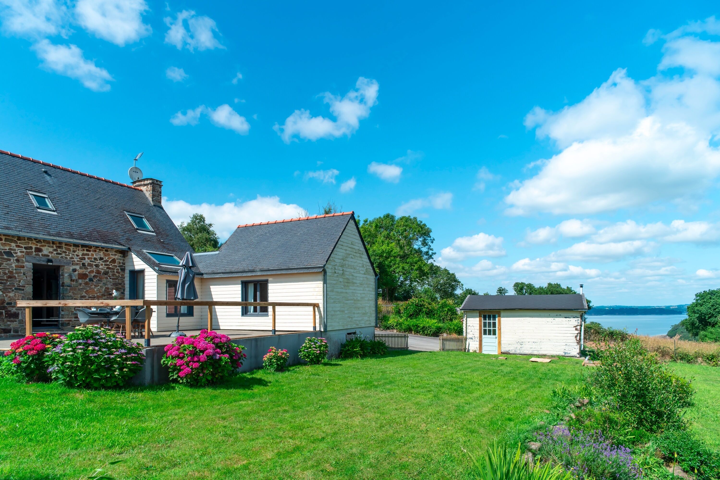 Une maison à deux étages avec une façade en pierre et un extérieur blanc, un petit abri séparé et une pelouse bien entretenue agrémentée de plantes à fleurs.