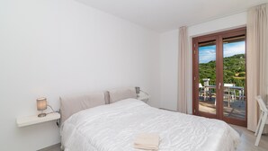 A bedroom with a bed, a bedside table with a lamp, a chair, and a balcony with a table and chairs.
