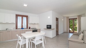 A modern kitchen with white cabinets, a dining table with chairs, a TV on a stand, and a sofa in a living area with a view of greenery outside.