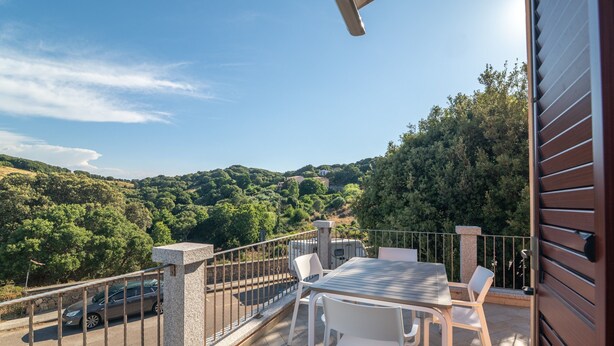 A balcony with a table and chairs, a stone pillar, and a view of a forested hillside.