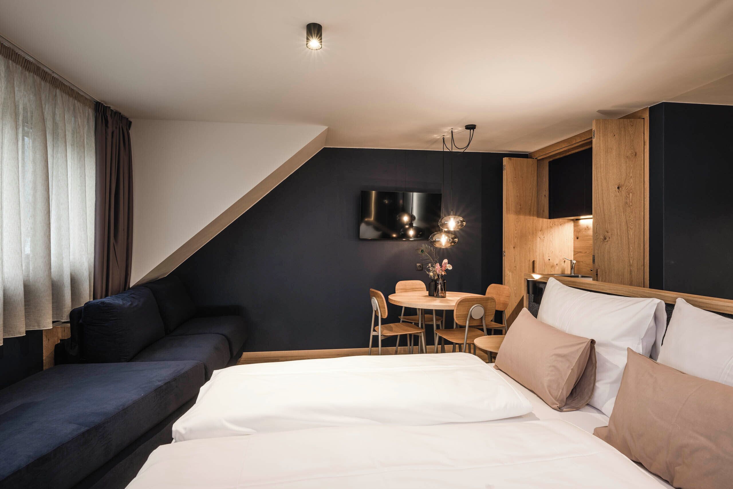 Une chambre d’hôtel moderne comprenant un lit, un canapé, une table à manger avec des chaises, un téléviseur fixé au mur et un meuble en bois.