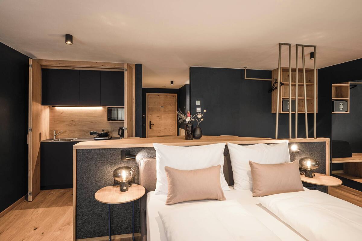 Ein modernes Hotelzimmer mit einer Küchenzeile, einem Bett mit zwei Kissen, Nachttischen mit Lampen und einer Holzleiter.