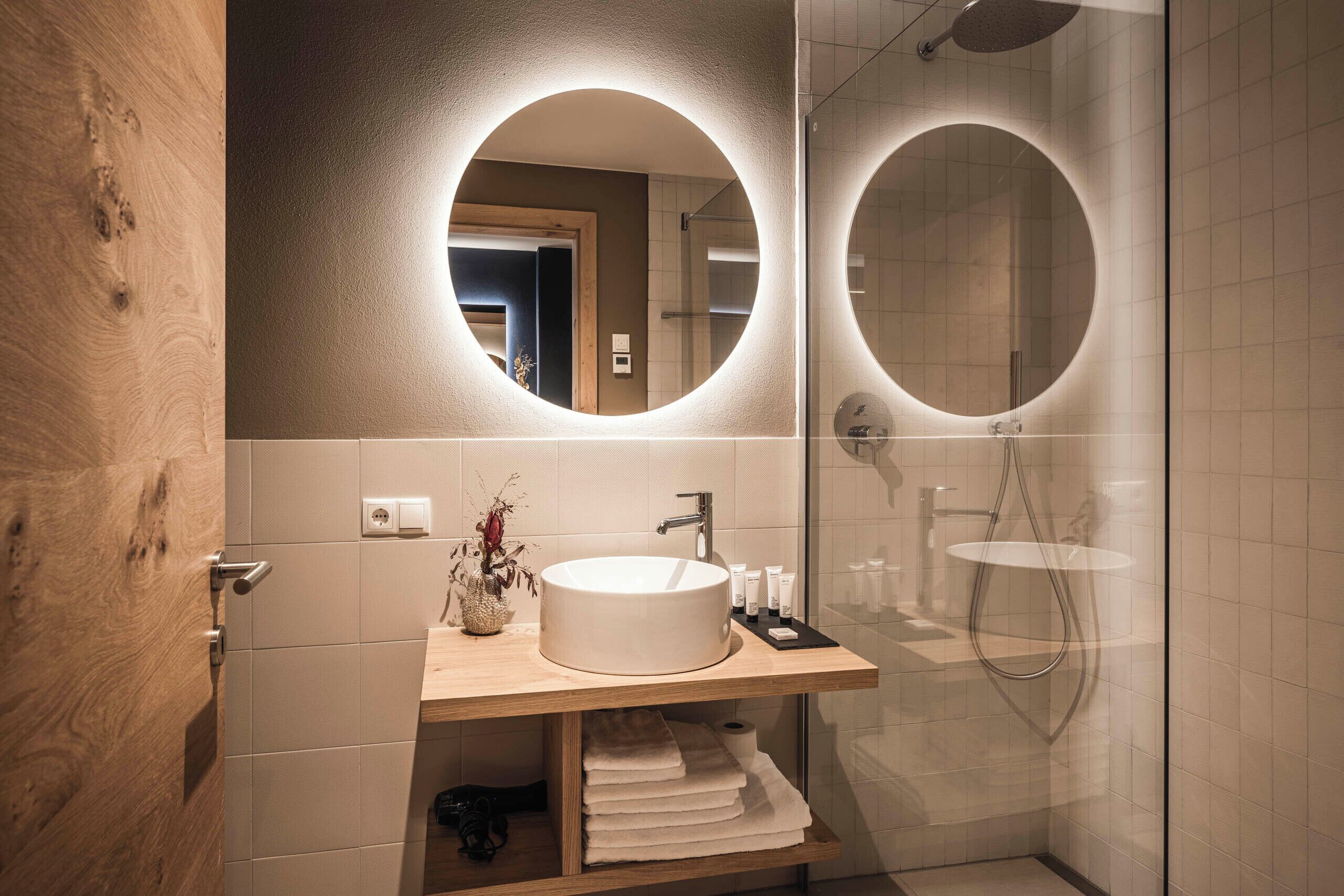 A modern bathroom with a wooden vanity, a round mirror, and a glass shower enclosure.