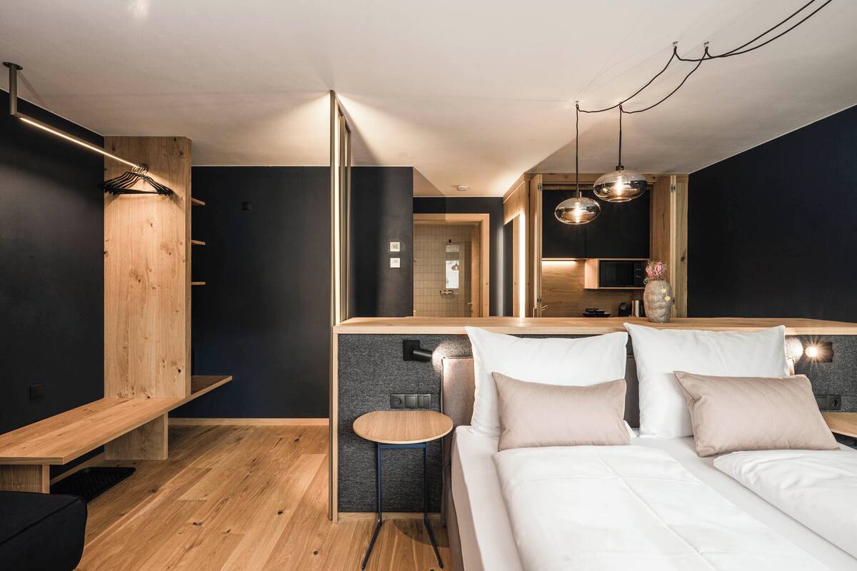 Ein modernes Hotelzimmer mit einer Wandpaneele aus Holz, einem kleinen runden Tisch, einem Bett mit weißer Bettwäsche und einer Küchenzeile.