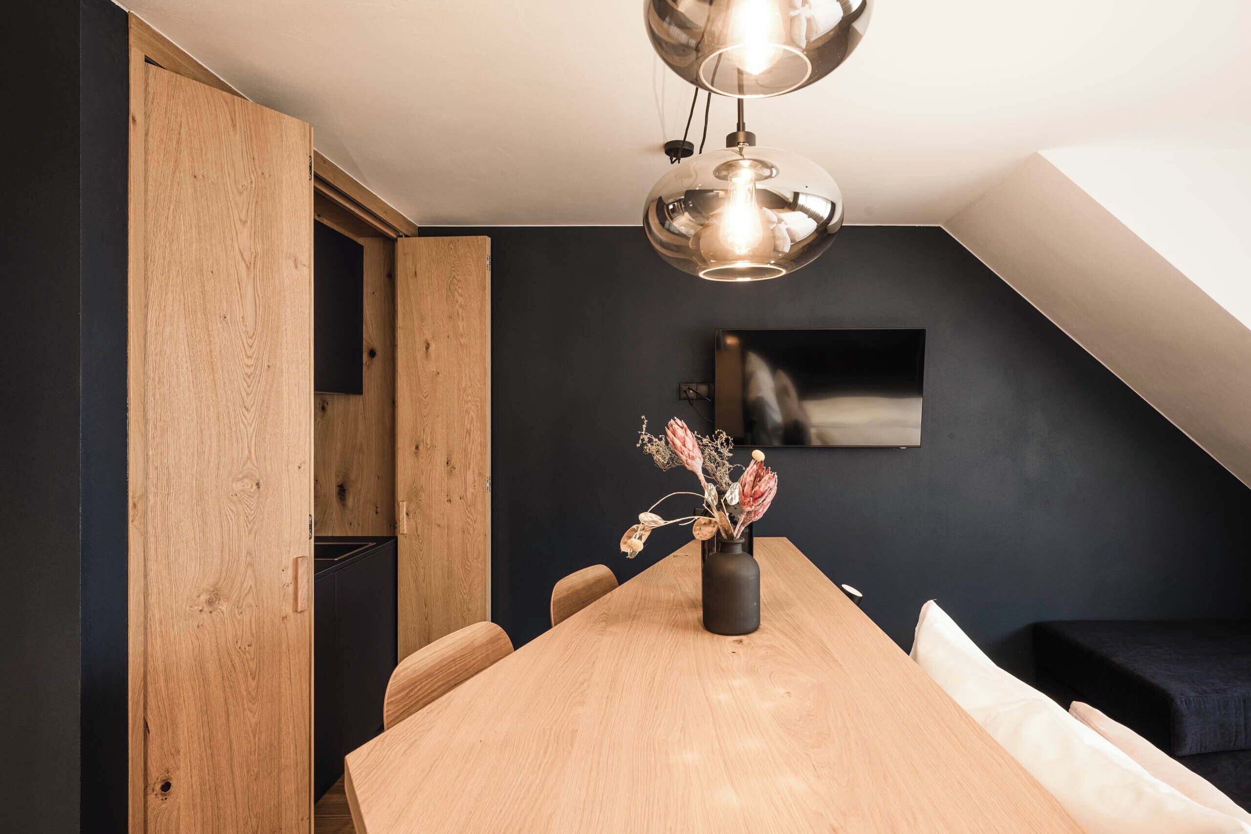 A modern dining area with a wooden table, chairs, a vase with dried flowers, a hanging light fixture, and a dark wall with a mounted TV.