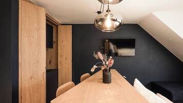 A modern dining area with a wooden table, chairs, a vase with dried flowers, a hanging light fixture, and a dark wall with a mounted TV.