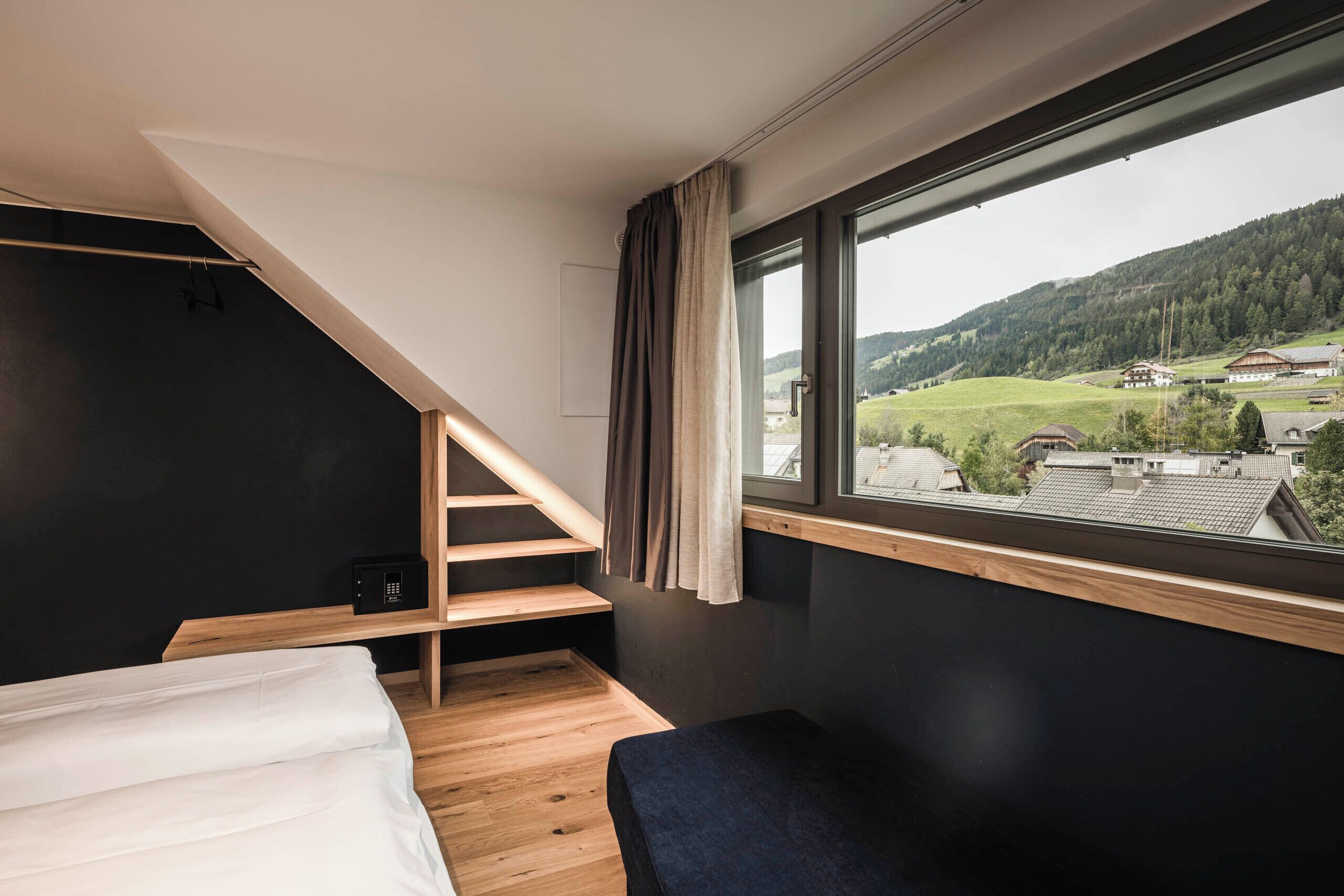 A bedroom with a bed, a bench, a small table, a window with a view of a valley, and a staircase.