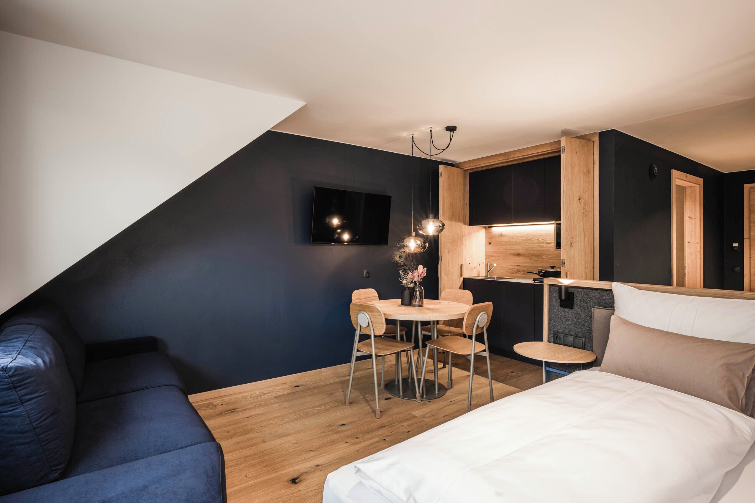Un dormitorio moderno y compacto con cama, una pequeña mesa redonda con sillas, televisor y una zona de kitchenette.