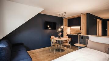 Un dormitorio moderno y compacto con cama, una pequeña mesa redonda con sillas, televisor y una zona de kitchenette.