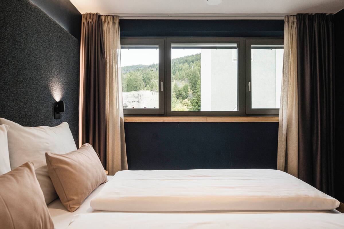 Ein Hotelzimmer mit Bett, Nachttisch, Wandlampe und einem Fenster mit Vorhängen.