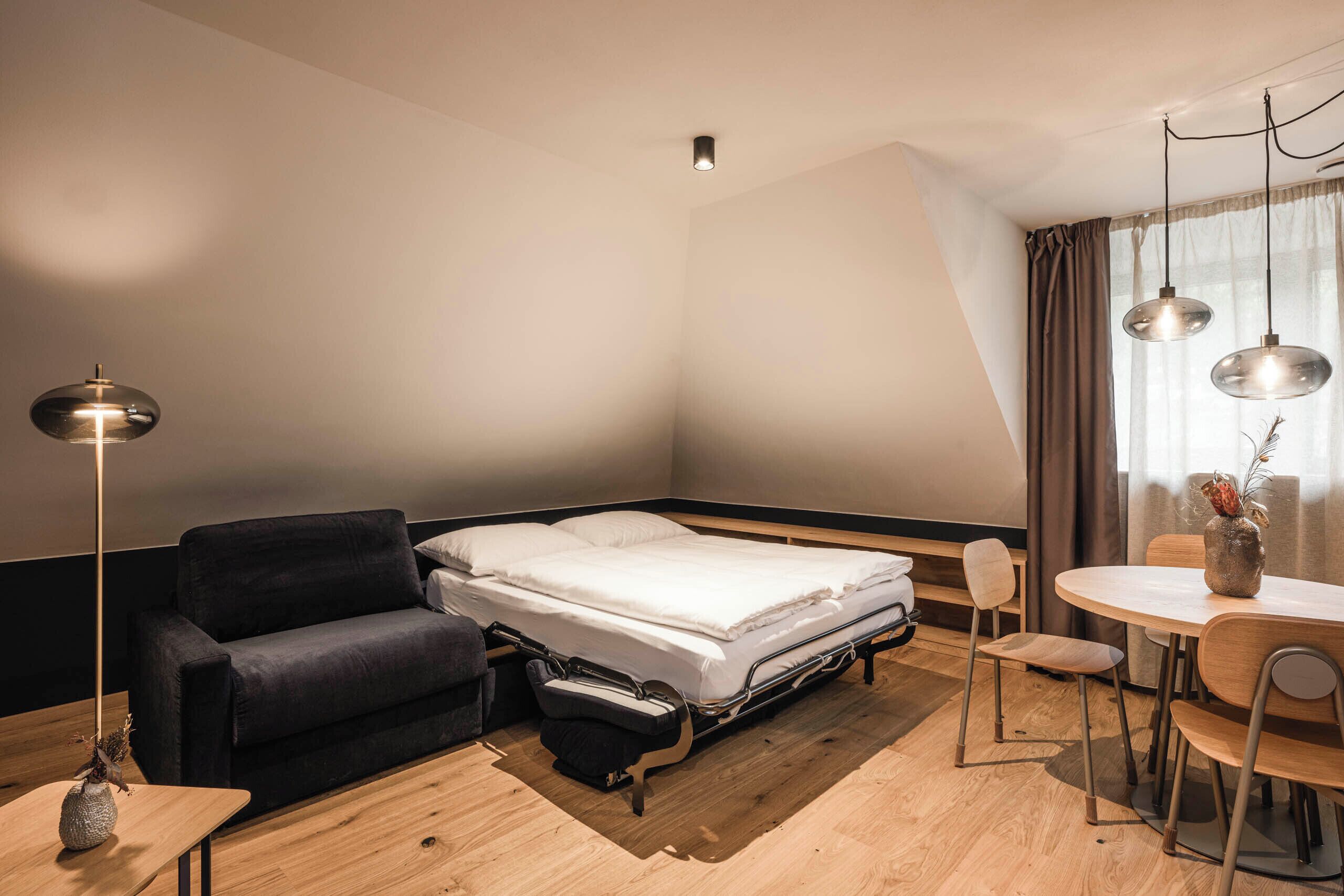 A compact hotel room with a bed, a sofa, a dining table, and a lamp.