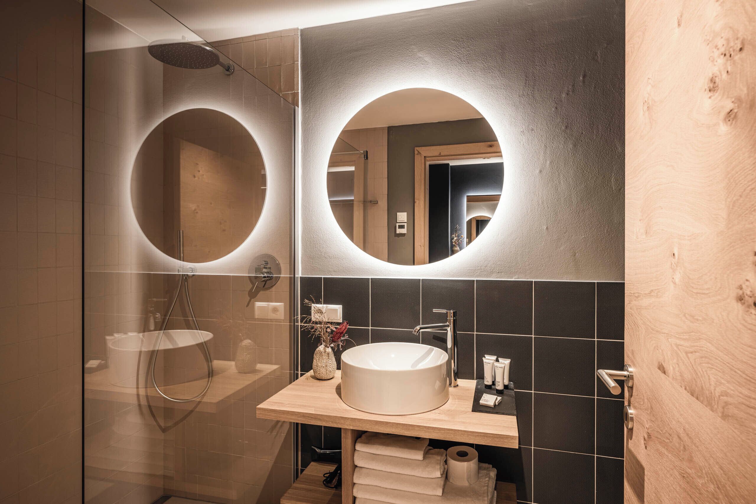 A modern bathroom with a glass-enclosed shower, a round mirror, and a wooden vanity with a sink.