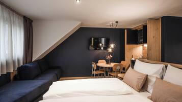 A modern hotel room with a bed, a sofa, a dining table with chairs, a TV mounted on the wall, and a wooden cabinet.