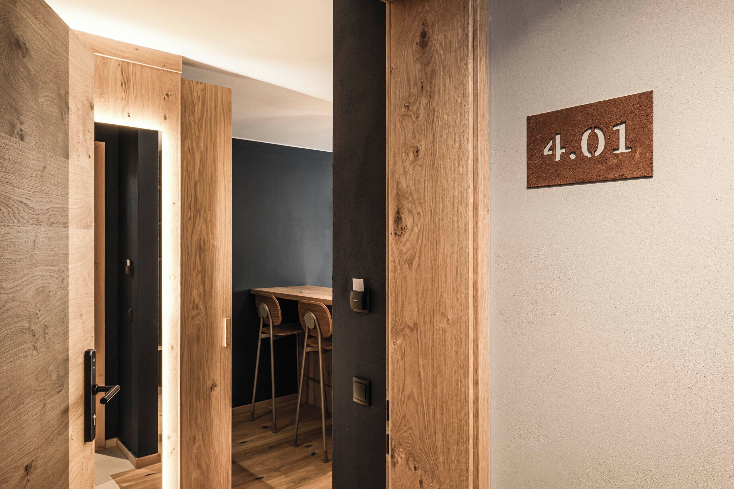 A hotel room with a wooden door, a small desk with two chairs, and a wall-mounted number plate reading "4.01".